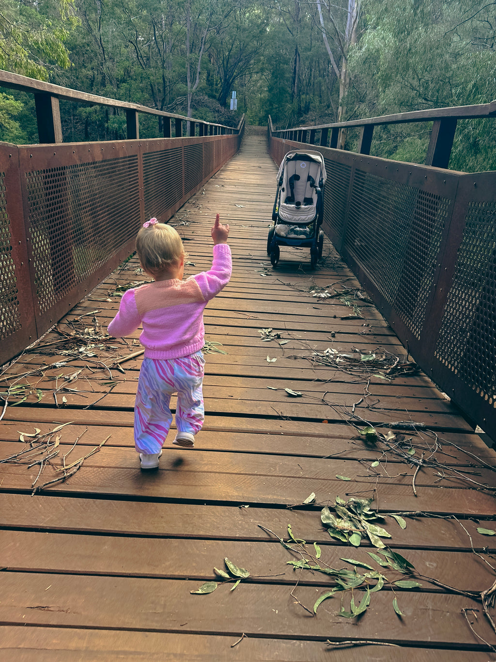 A toddler in a pink outfit walks up a wooden boardwalk bridge toward a stroller, with trees and foliage visible in the background.