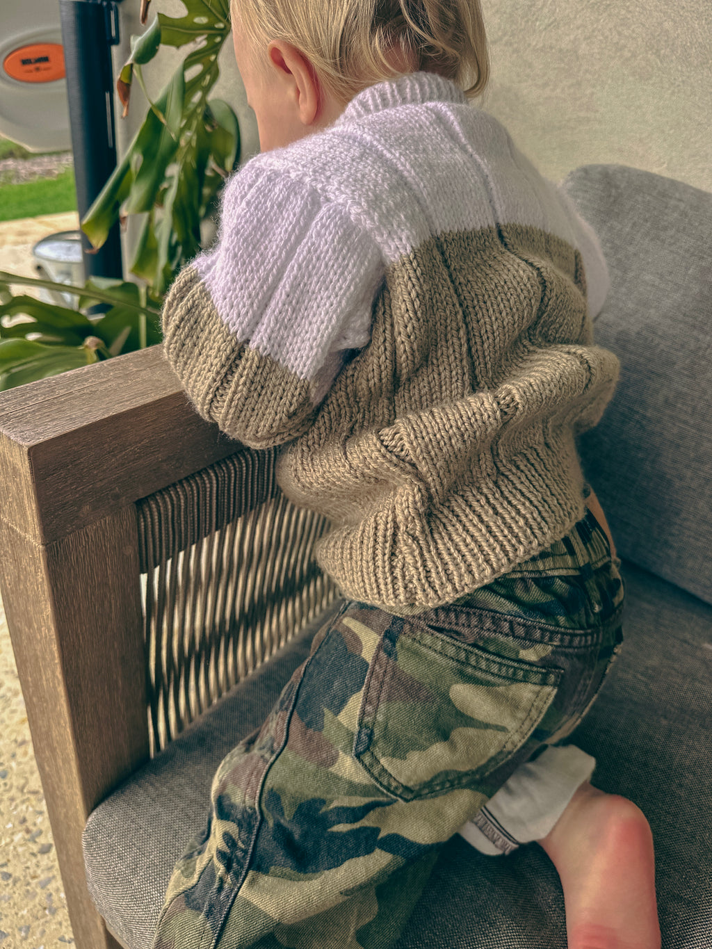 A child wearing a chunky knit color-block sweater in white and beige, paired with camouflage pants, sitting on a gray outdoor sofa.