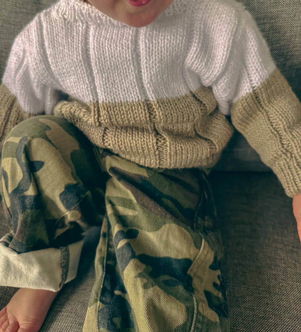 A close-up of a child wearing a chunky knit color-block sweater with a white upper section and beige sleeves & lower body, paired with camouflage pants, sitting on a gray textured couch.