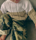 A close-up of a child wearing a chunky knit color-block sweater with a white upper section and beige sleeves & lower body, paired with camouflage pants, sitting on a gray textured couch.