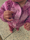 A close-up of a child wearing a chunky hand-knit cardigan in a variegated pink yarn, standing on a speckled stone tile floor.
