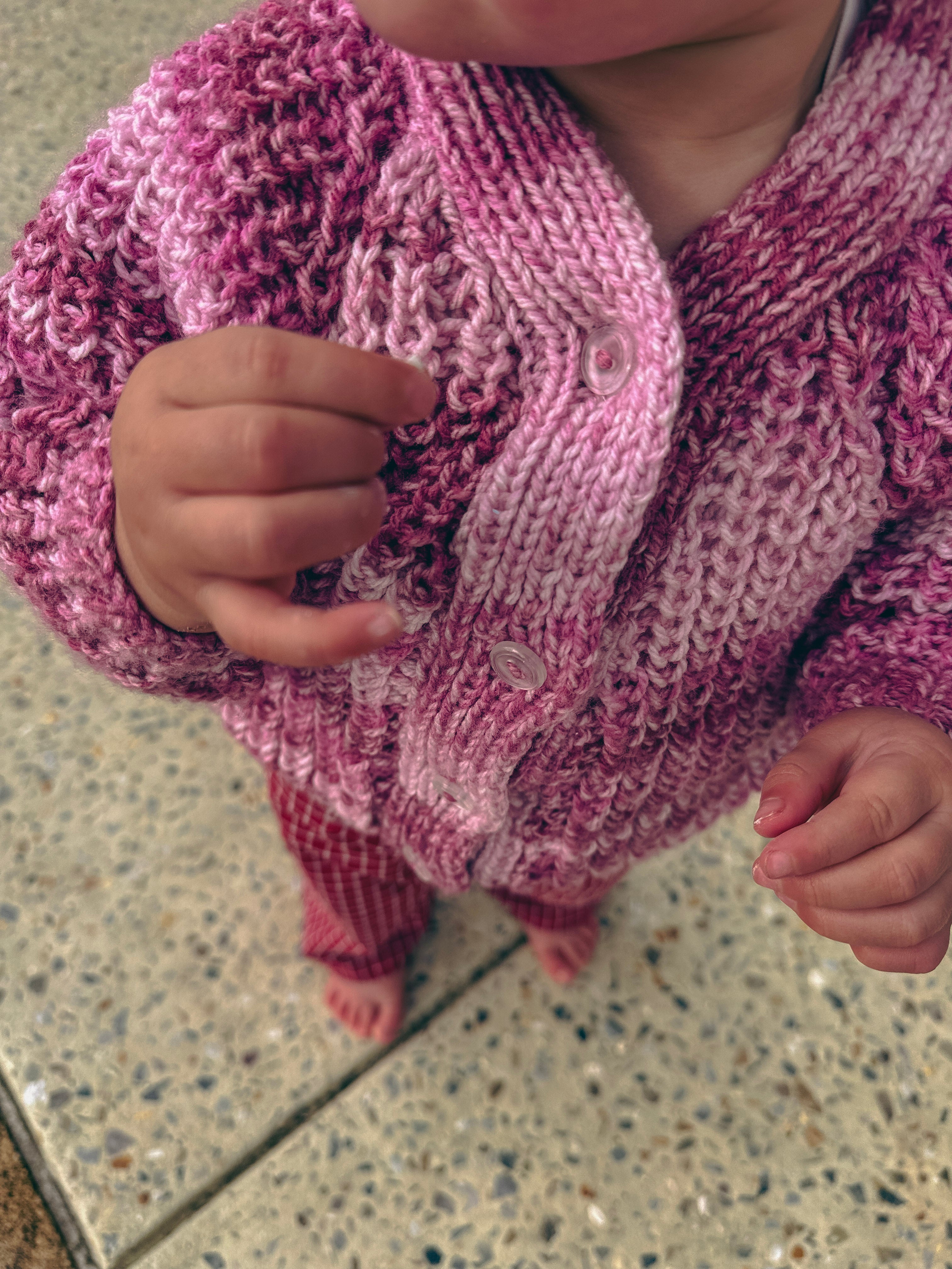A close-up of a child wearing a chunky hand-knit cardigan in a variegated pink yarn, standing on a speckled stone tile floor.