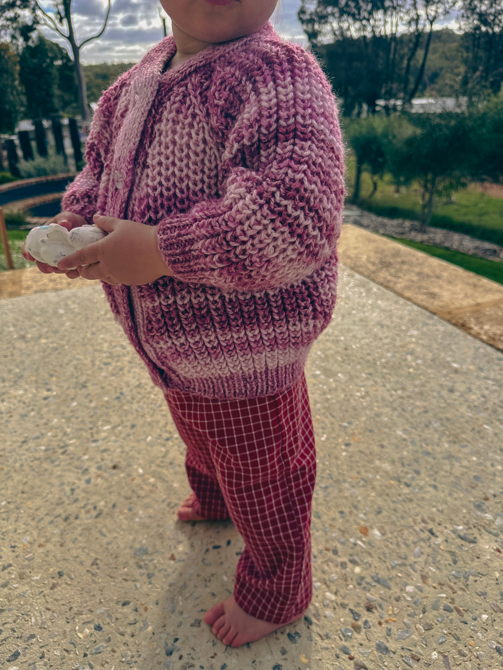 A child standing outdoors on a paved path wearing a chunky knit zip-up cardigan in pink and white, paired with matching pink plaid pants.