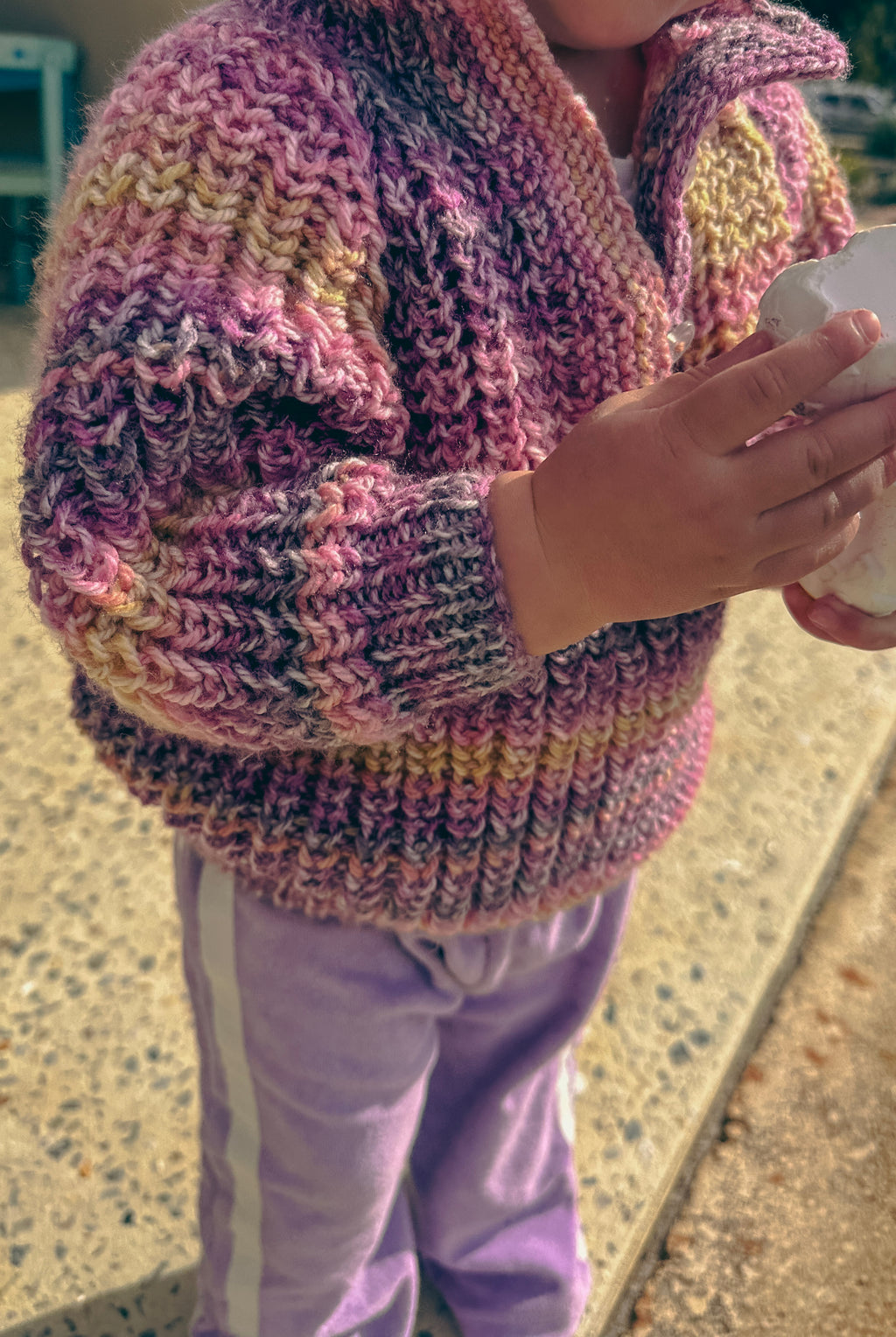 A close-up of a child wearing a chunky hand-knit cardigan sweater in a variegated pink, purple, and yellow colorway, paired with lavender pants.