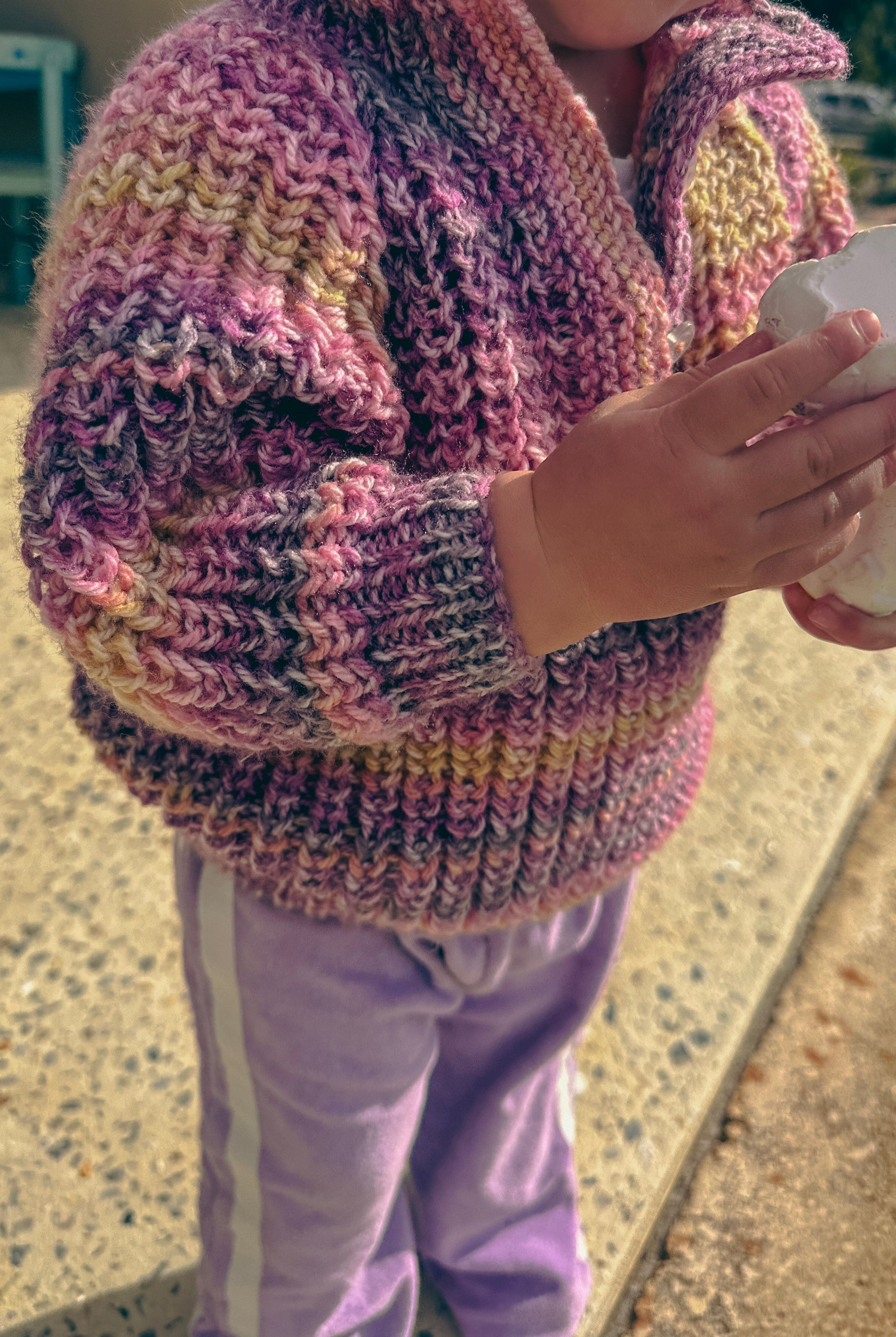 A close-up of a child wearing a chunky hand-knit cardigan sweater in a variegated pink, purple, and yellow colorway, paired with lavender pants.