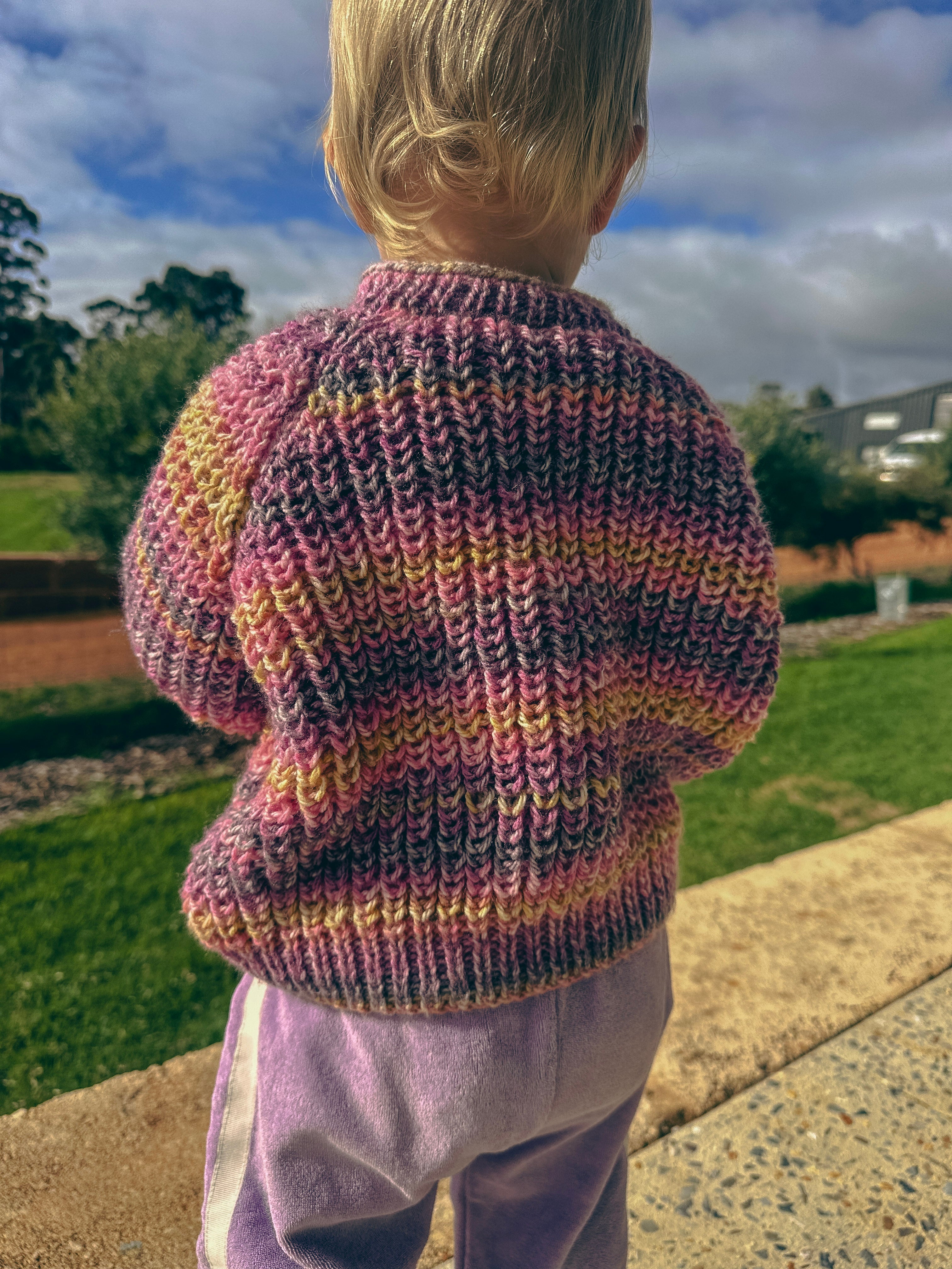 A child viewed from behind wearing a hand-knit cardigan sweater with horizontal stripes of pink, purple, yellow, and gray, paired with light purple pants, standing outdoors on a sunny day.