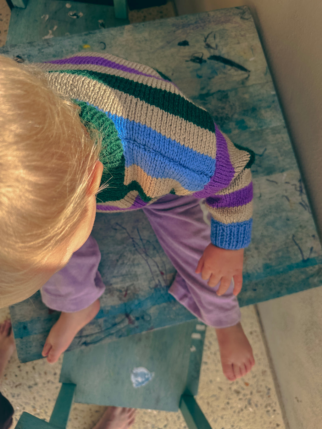 A child wearing a colourful striped knit sweater and purple pants sits on a worn wooden stool in an outdoor setting.
