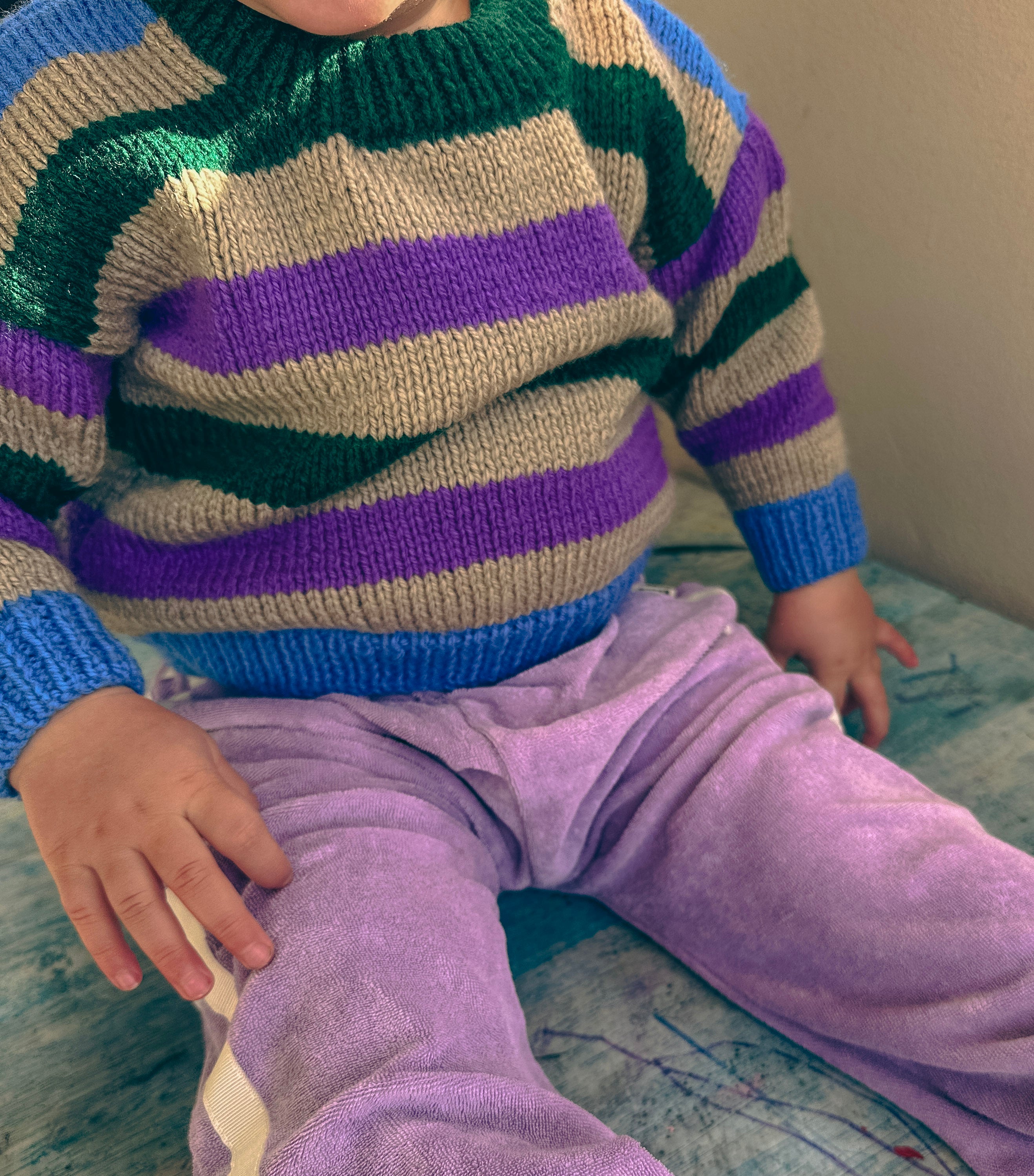 A close-up of a child wearing a hand-knit crewneck sweater with horizontal stripes of green, purple, grey, and blue, paired with light purple pants.