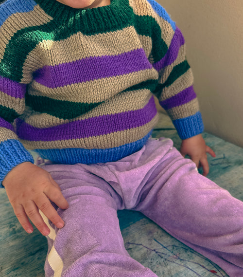 A close-up of a child wearing a hand-knit crewneck sweater with horizontal stripes of green, purple, grey, and blue, paired with light purple pants.