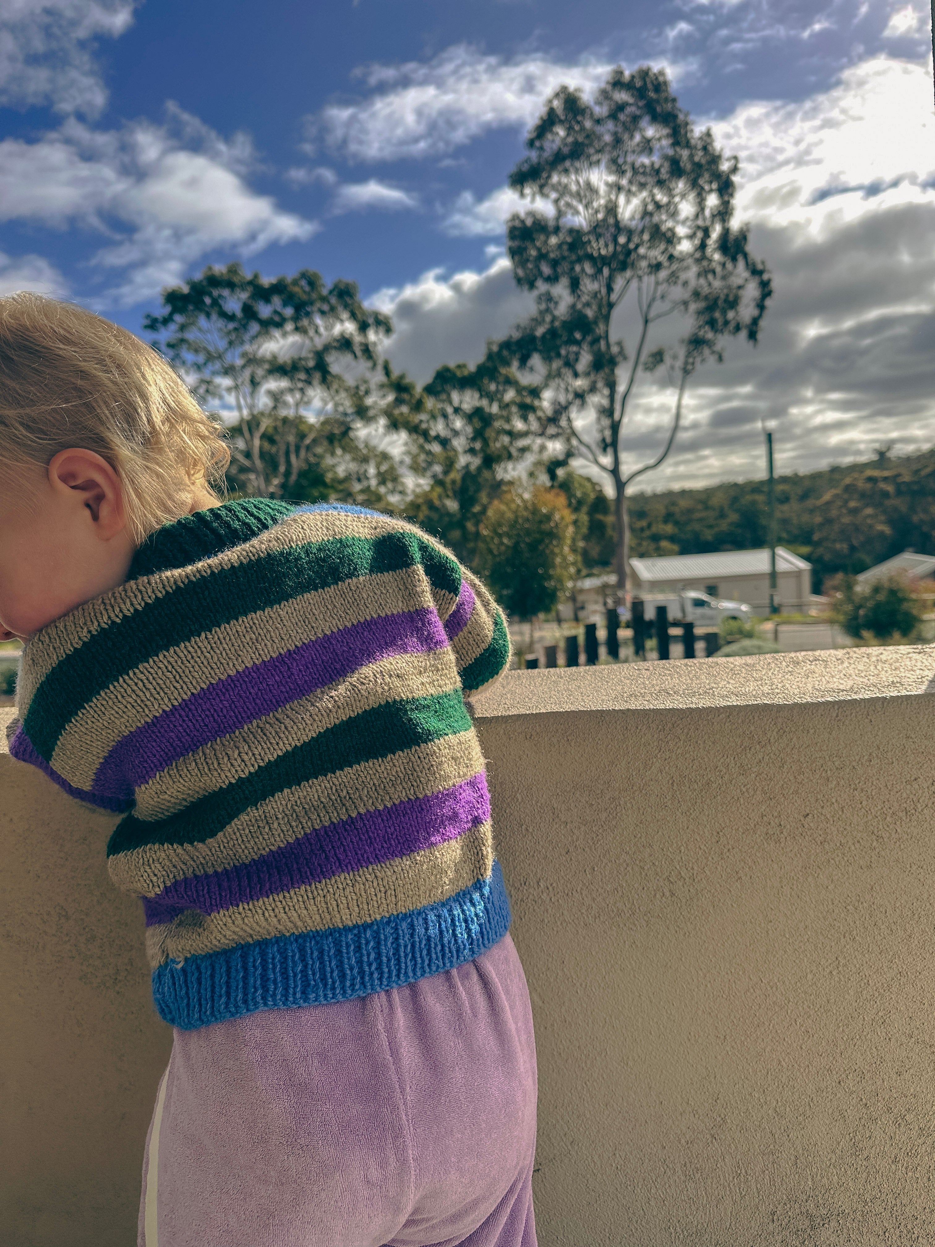 A child wearing a striped knit sweater with green, purple, grey, and blue horizontal stripes, standing outdoors against a beige wall with trees and a blue sky in the background.