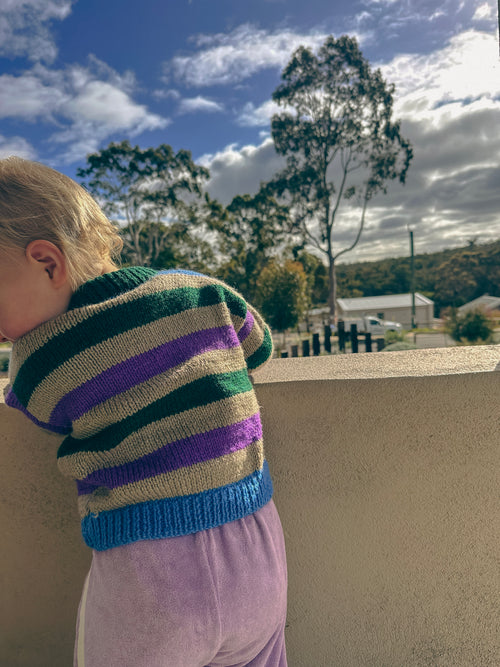 A child wearing a striped knit sweater with green, purple, grey, and blue horizontal stripes, standing outdoors against a beige wall with trees and a blue sky in the background.