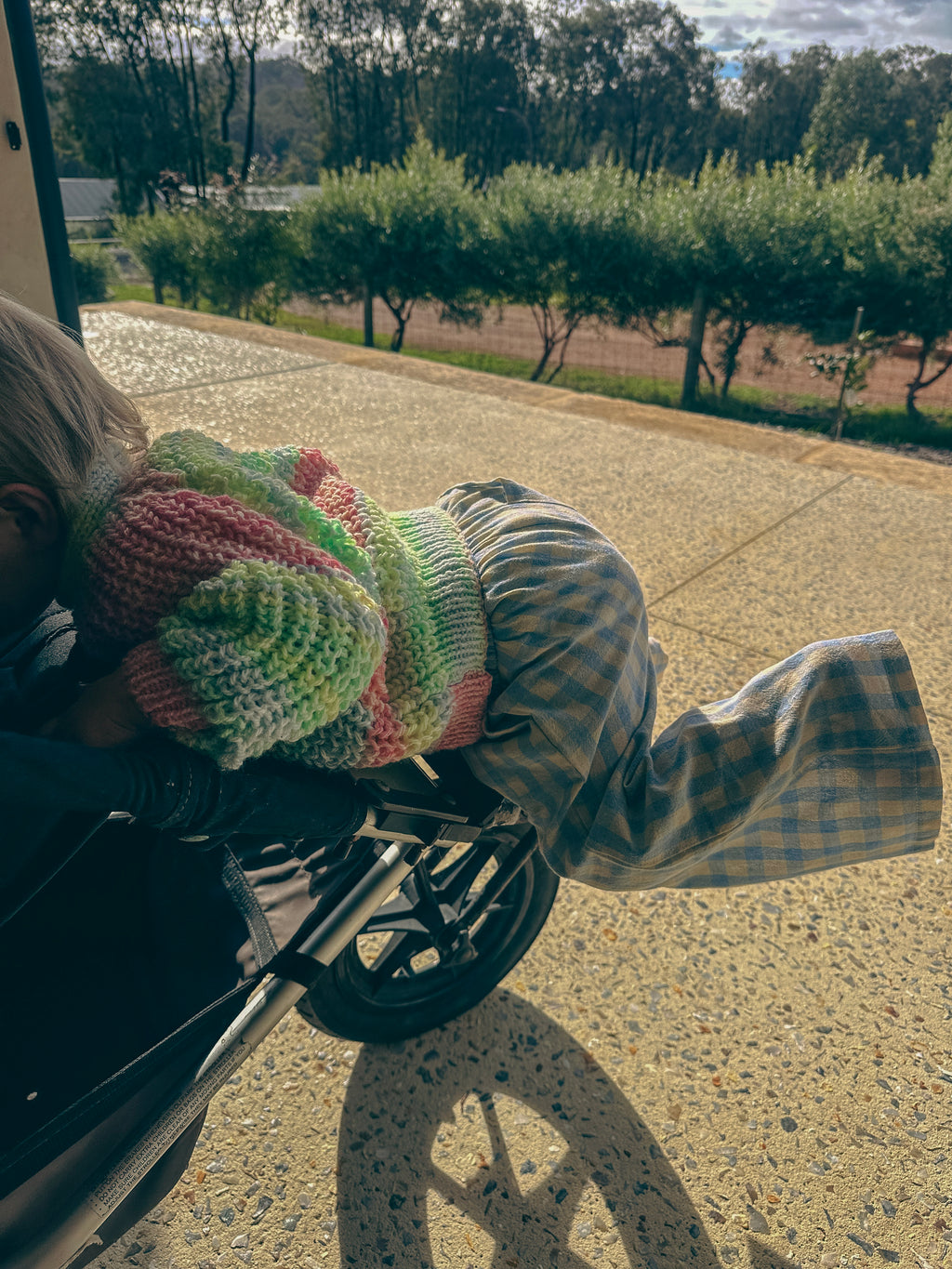 A child wearing a colorful striped knit hat and plaid pants leans over a stroller outdoors on a paved driveway with trees in the background.