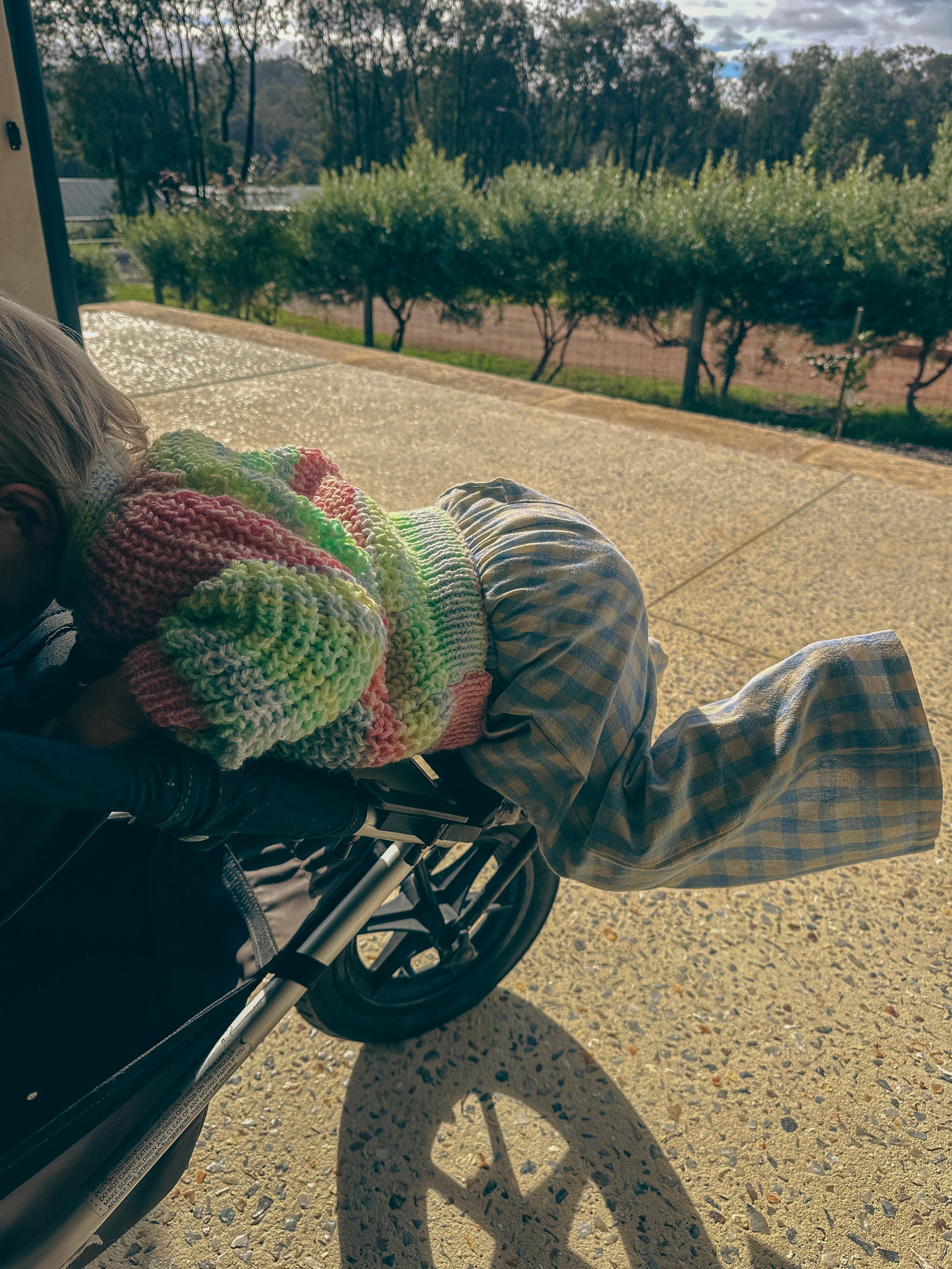 A child wearing a colorful striped knit hat and plaid pants leans over a stroller outdoors on a paved driveway with trees in the background.
