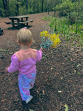 A toddler seen from behind wearing a pink and purple knit sweater and patterned pants, walking on a dirt path in a wooded outdoor setting while holding yellow flowers.