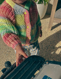 A close-up of a child wearing a colourful striped knit cardigan with pastel pink, green, yellow, and blue stripes, standing in a sunlit outdoor setting.
