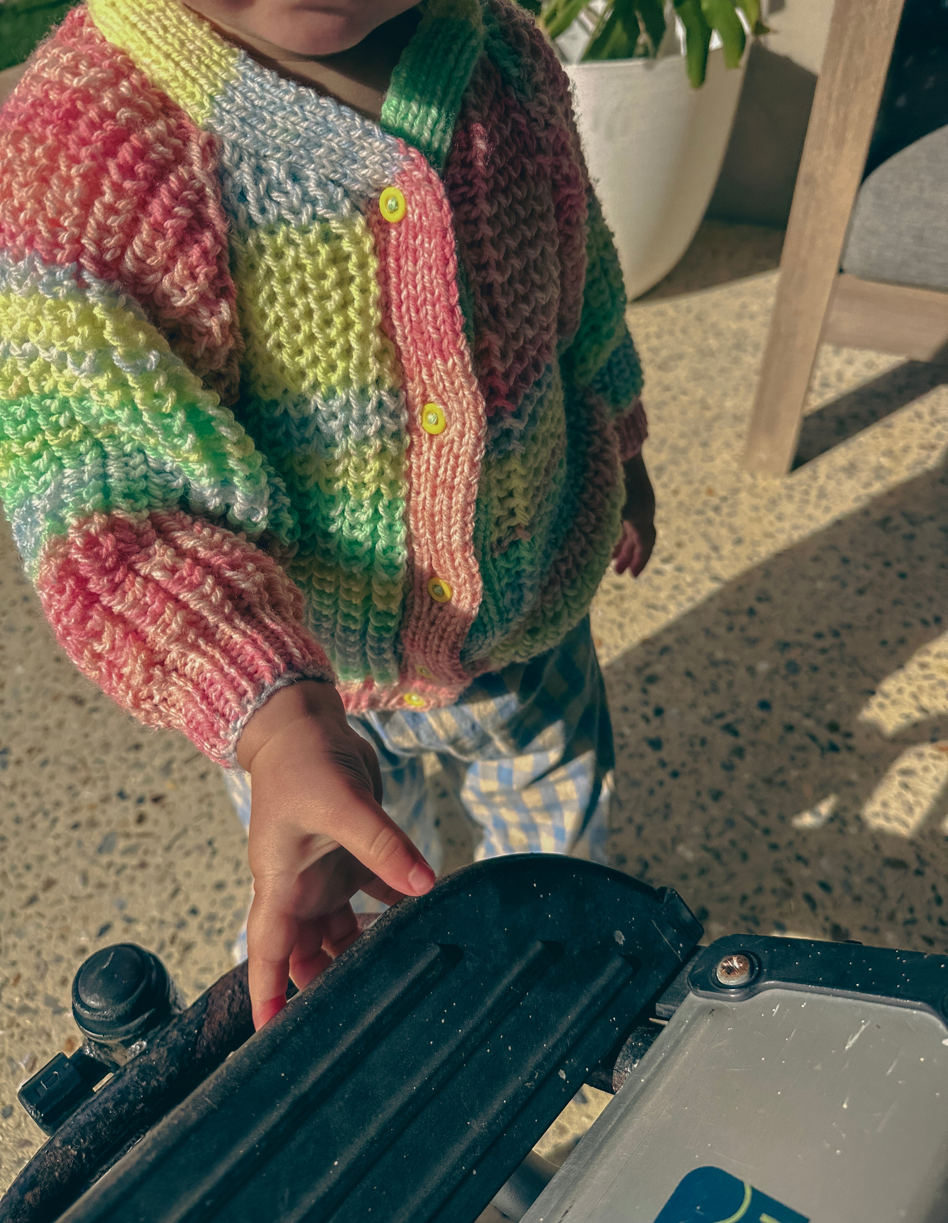 A close-up of a child wearing a colourful striped knit cardigan with pastel pink, green, yellow, and blue stripes, standing in a sunlit outdoor setting.