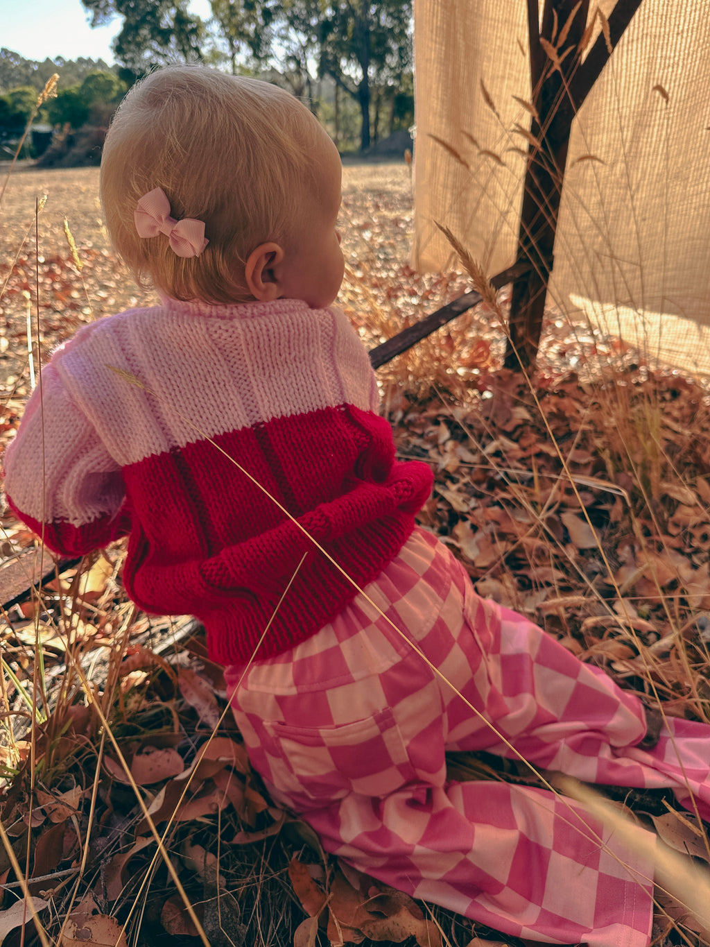 A baby sitting outdoors on dry leaves, wearing a pink and red knit top with pink and white checkered pants.