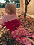 A baby sitting outdoors on dry leaves, wearing a pink and red knit top with pink and white checkered pants.