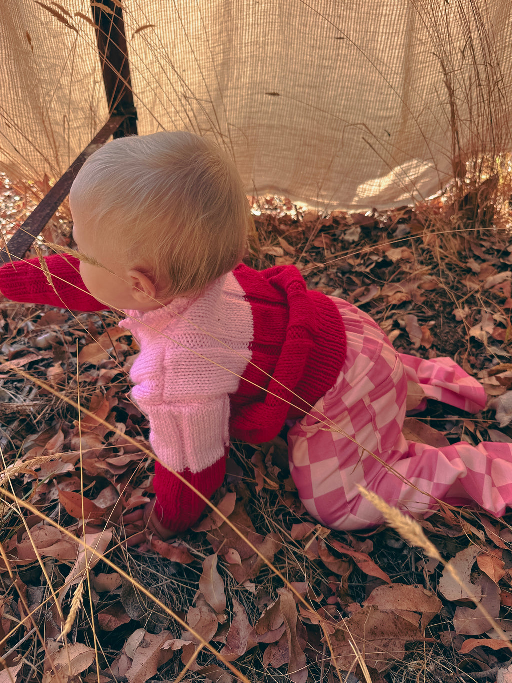 A baby wearing a pink and red knit hooded sweater and pink patterned pants crawls on dry leaves outdoors under a beige canvas cover.