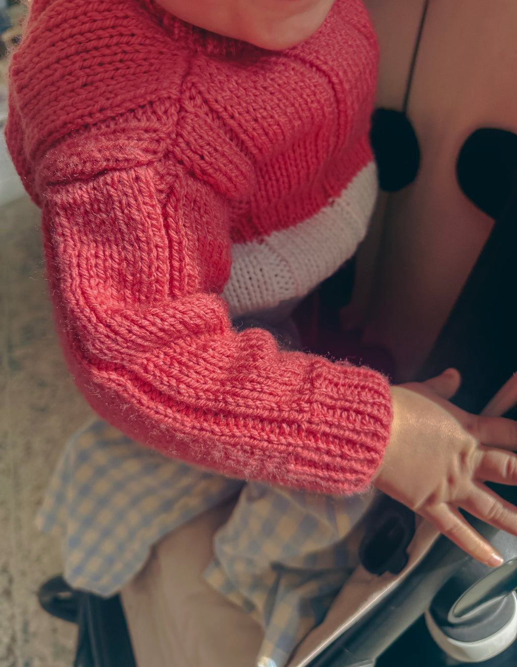 A close-up of a child wearing a chunky knit sweater with a coral pink body and a contrasting white band across the chest, seated in a stroller.
