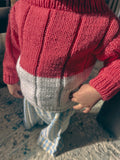 A close-up of a child wearing a chunky knit sweater with a color-block design, featuring a dark red upper section and a white lower section, paired with blue and white checkered trousers.