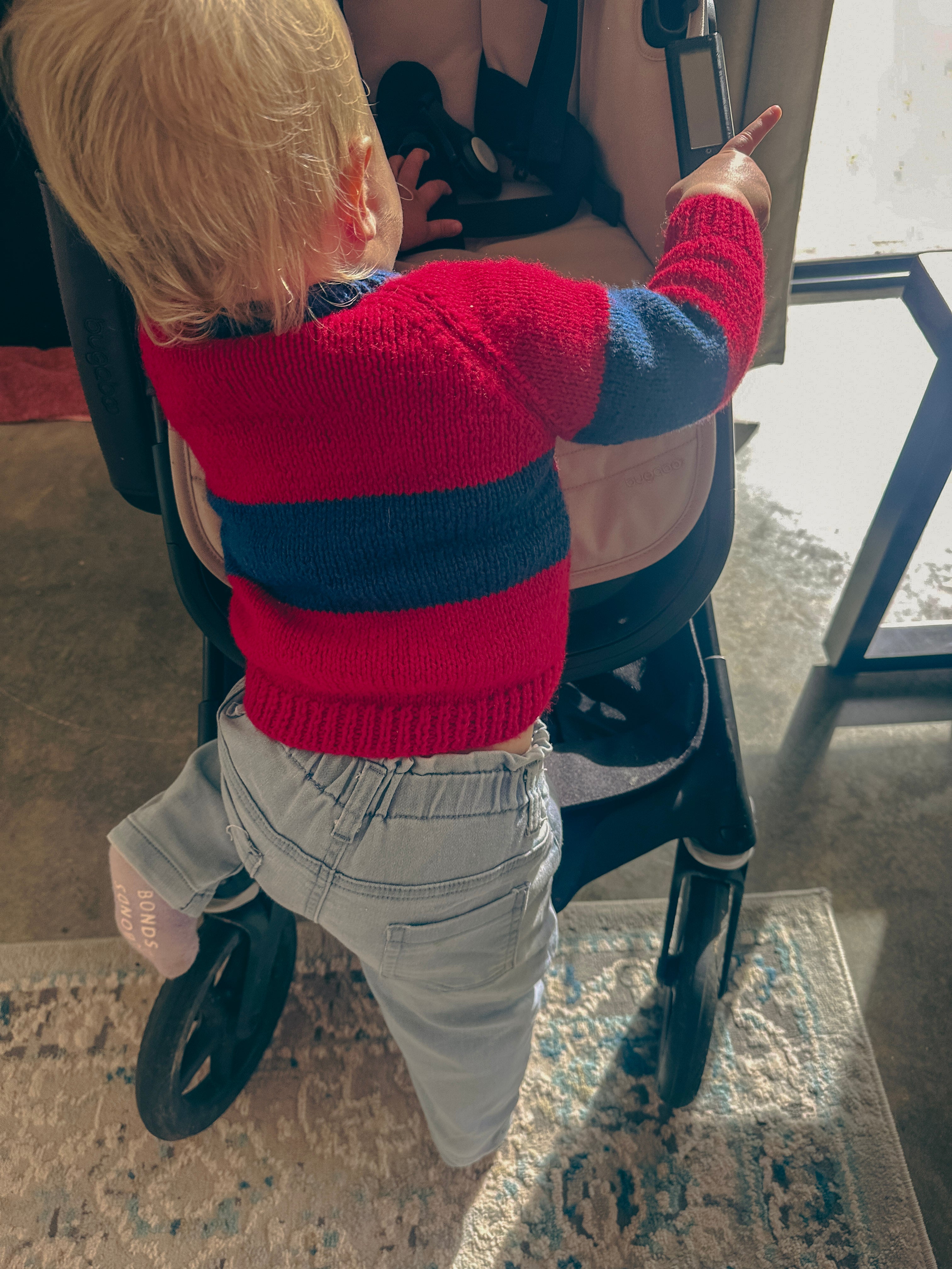 A toddler with blonde hair wearing a red and navy blue striped knit sweater and light gray pants, standing inside a stroller and pointing toward something outside the frame.