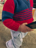 A close-up of a child wearing a red and navy blue striped knit sweater and light wash jeans, holding a black tablet in a home setting.