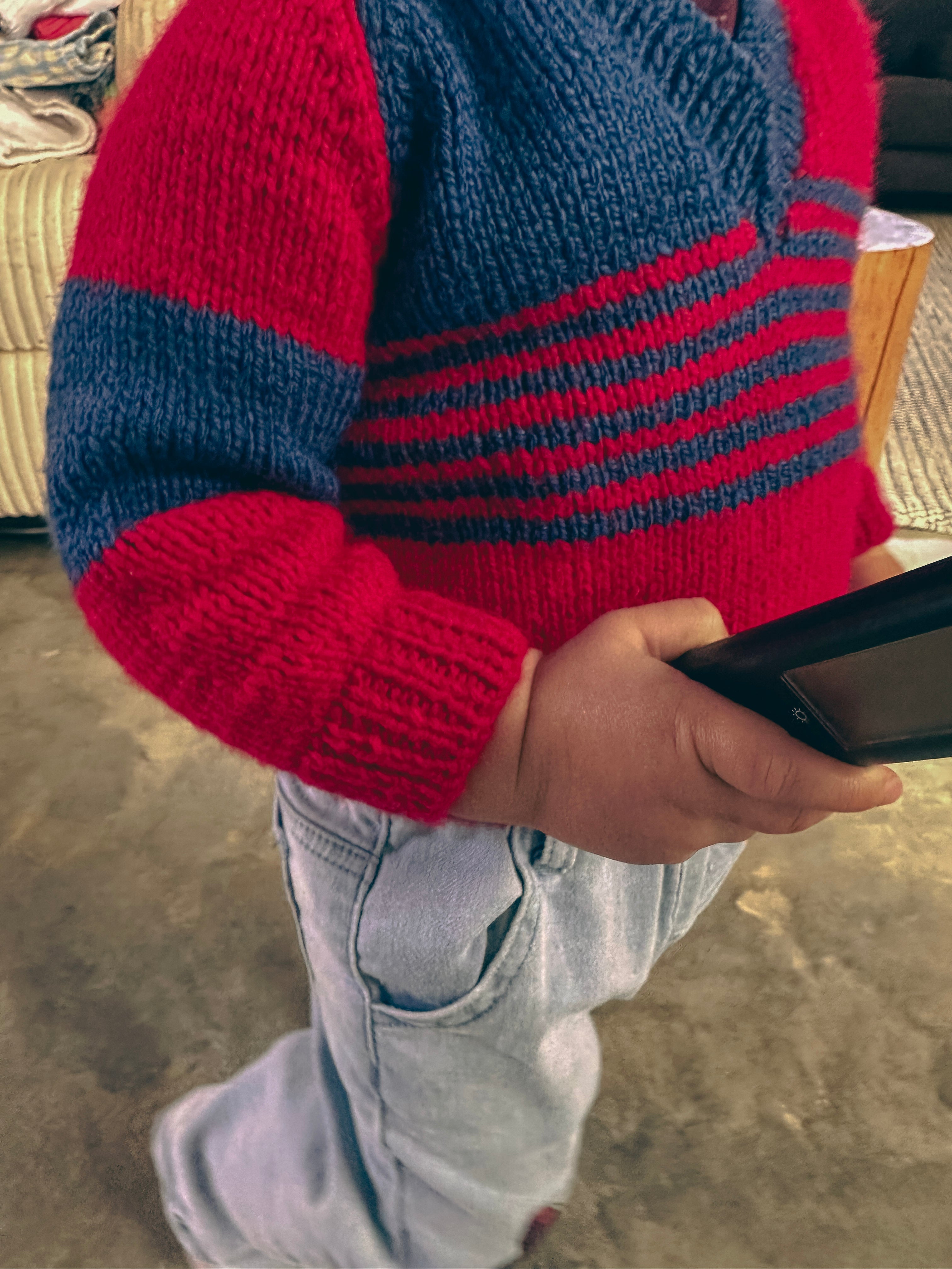 A close-up of a child wearing a red and navy blue striped knit sweater and light wash jeans, holding a black tablet in a home setting.