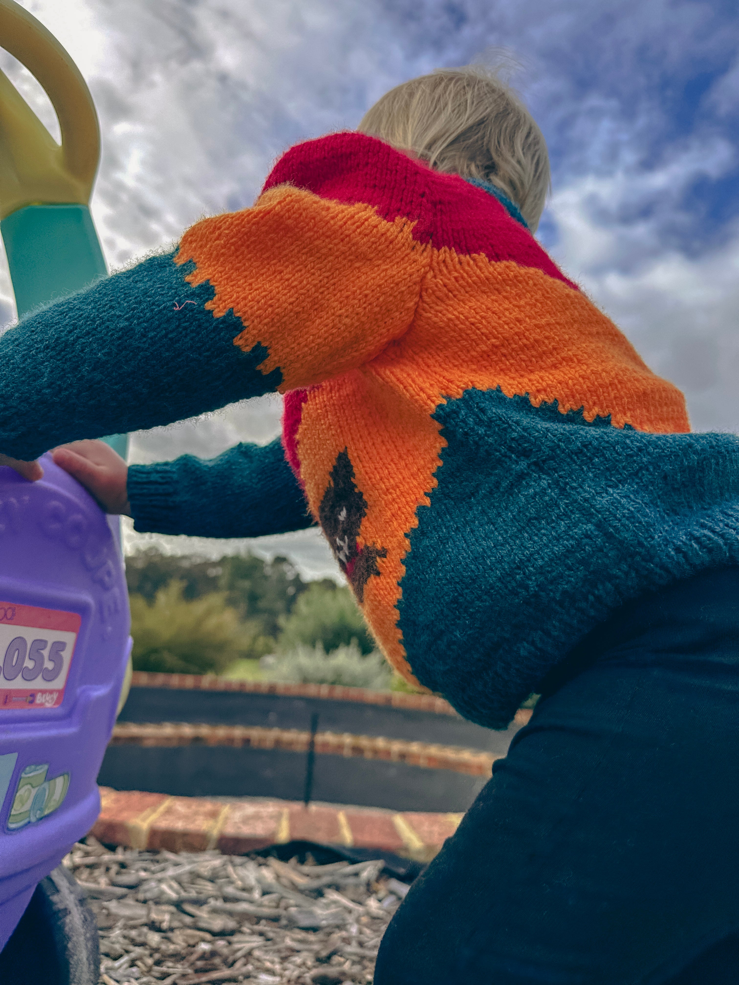 A child wearing a colorful hand-knit sweater with orange, teal, and red sections, seen from behind while playing outdoors on a playground.