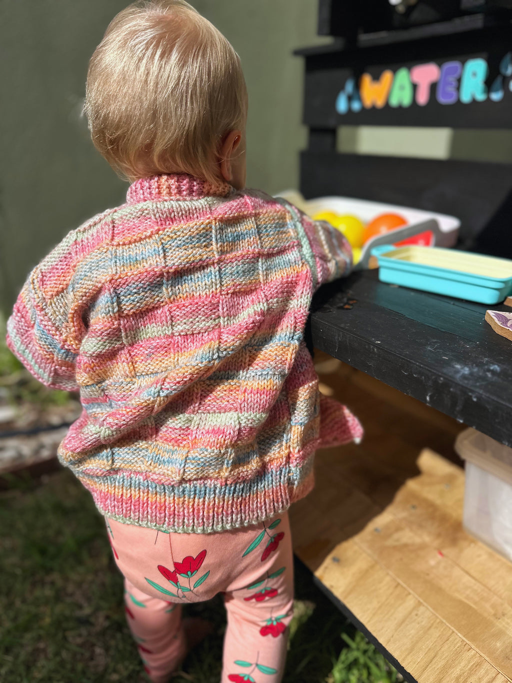Child wearing a colorful striped sweater and pink pants with floral designs, standing near a toy kitchen set.