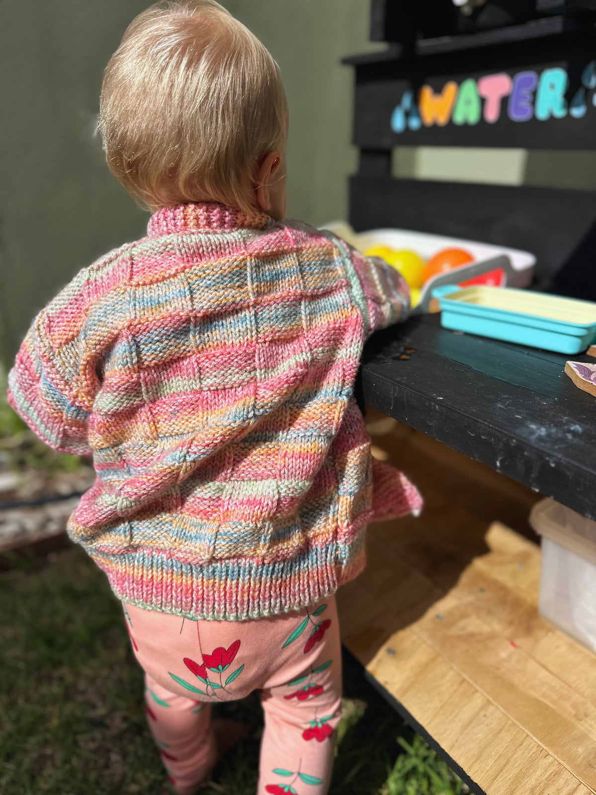 Child wearing a colorful striped sweater and pink pants with floral designs, standing near a toy kitchen set.
