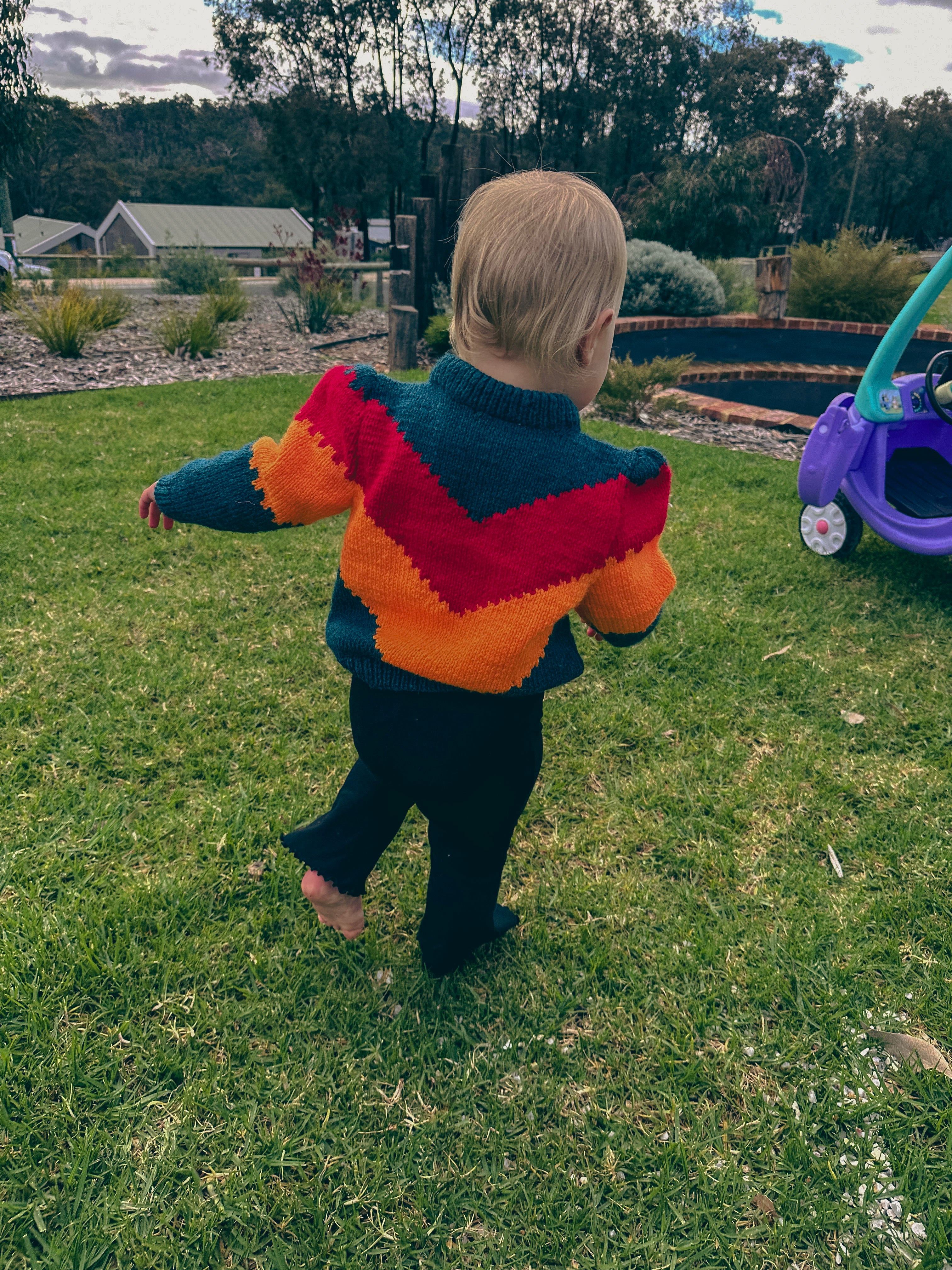 A toddler walking barefoot on a grassy backyard, wearing a colorful hand-knit sweater with a chevron pattern in teal, red, and orange, paired with black pants.