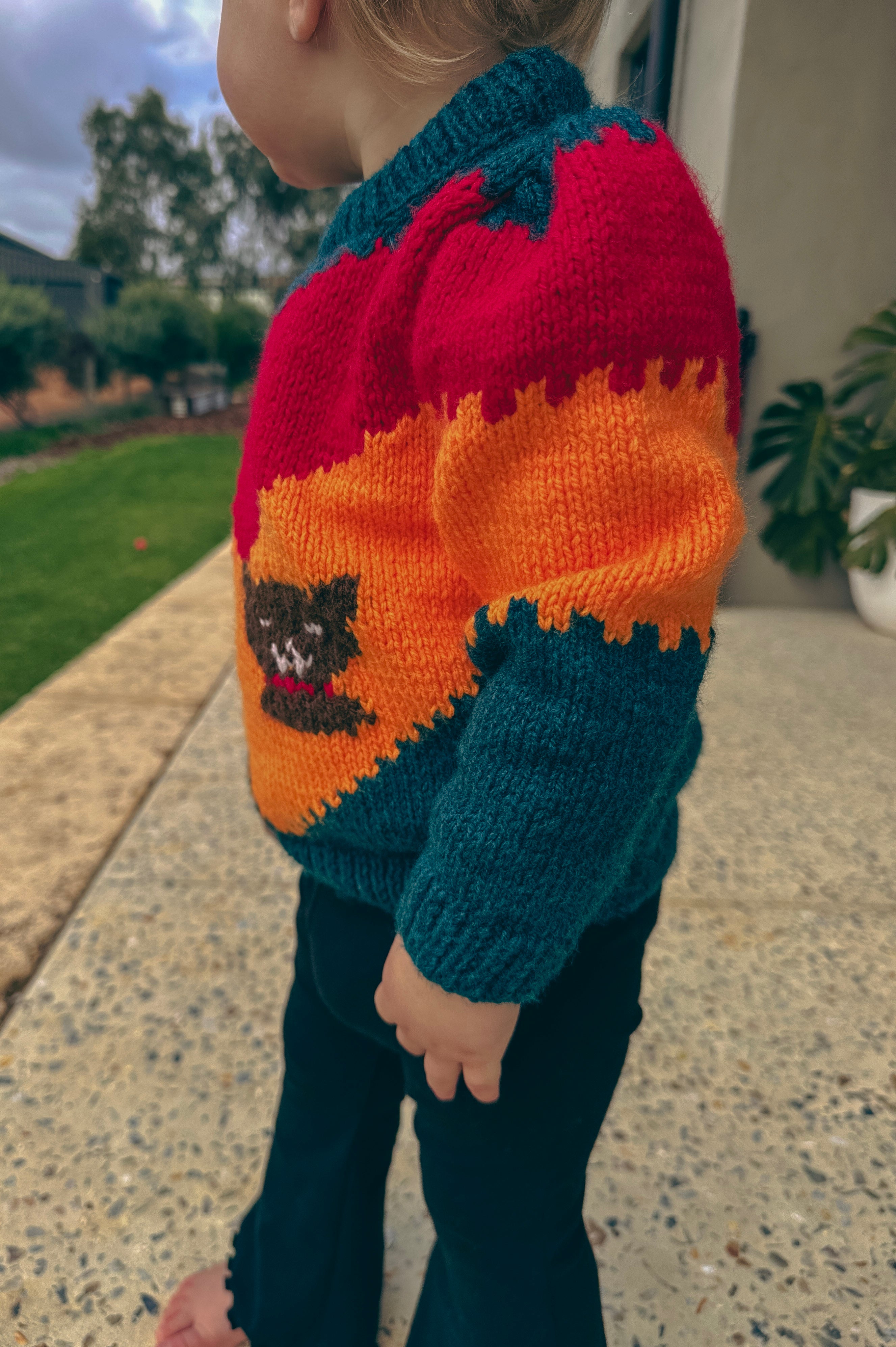 A toddler wearing a colorful hand-knit sweater with red, orange, and teal sections, featuring a crocheted dog design on the front, standing outdoors on a concrete patio.