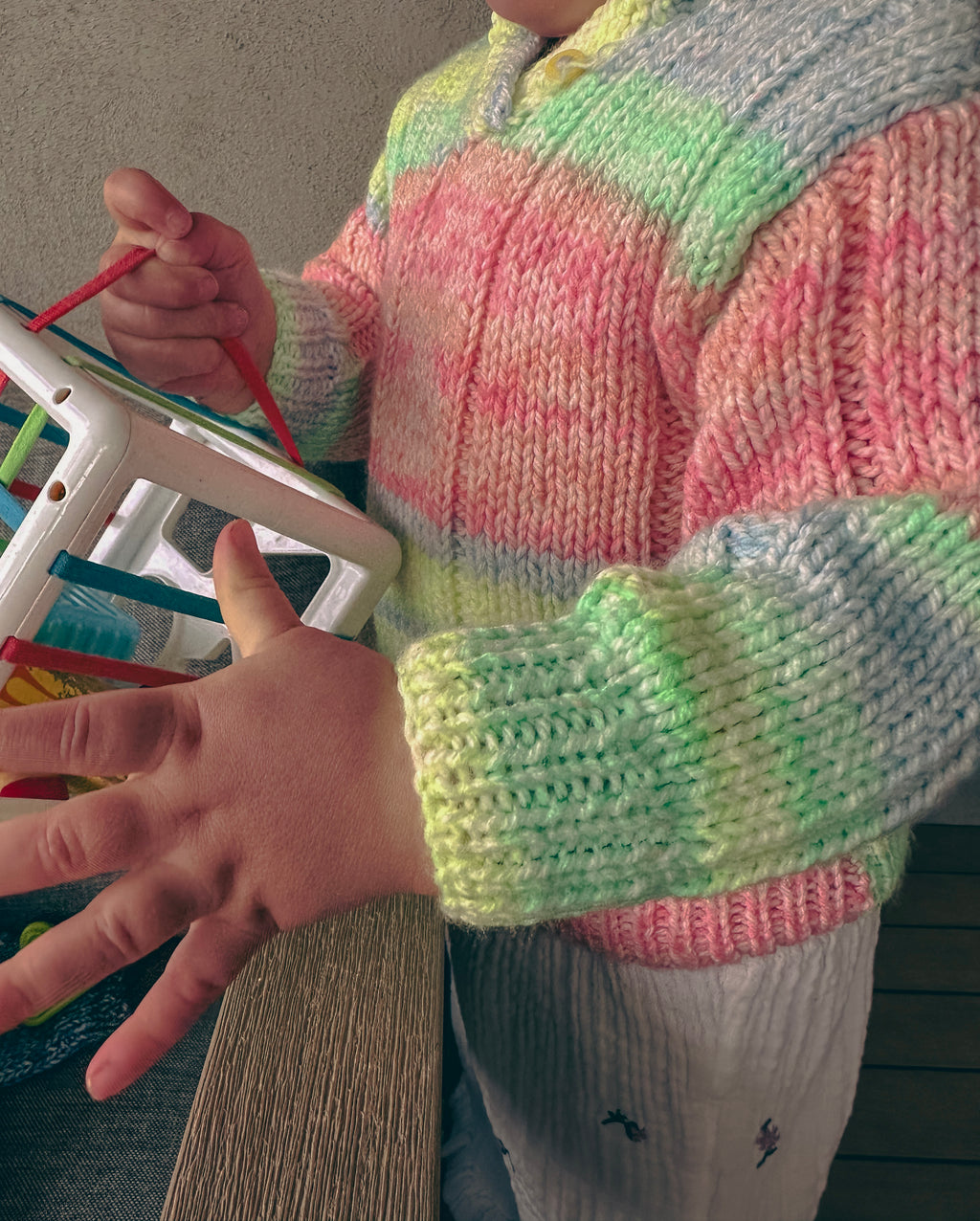 A close-up of a person wearing a chunky knit sweater with pastel rainbow stripes in pink, green, yellow, blue, and white. The child is holding a small white toy or frame in one hand while gesturing with the other.