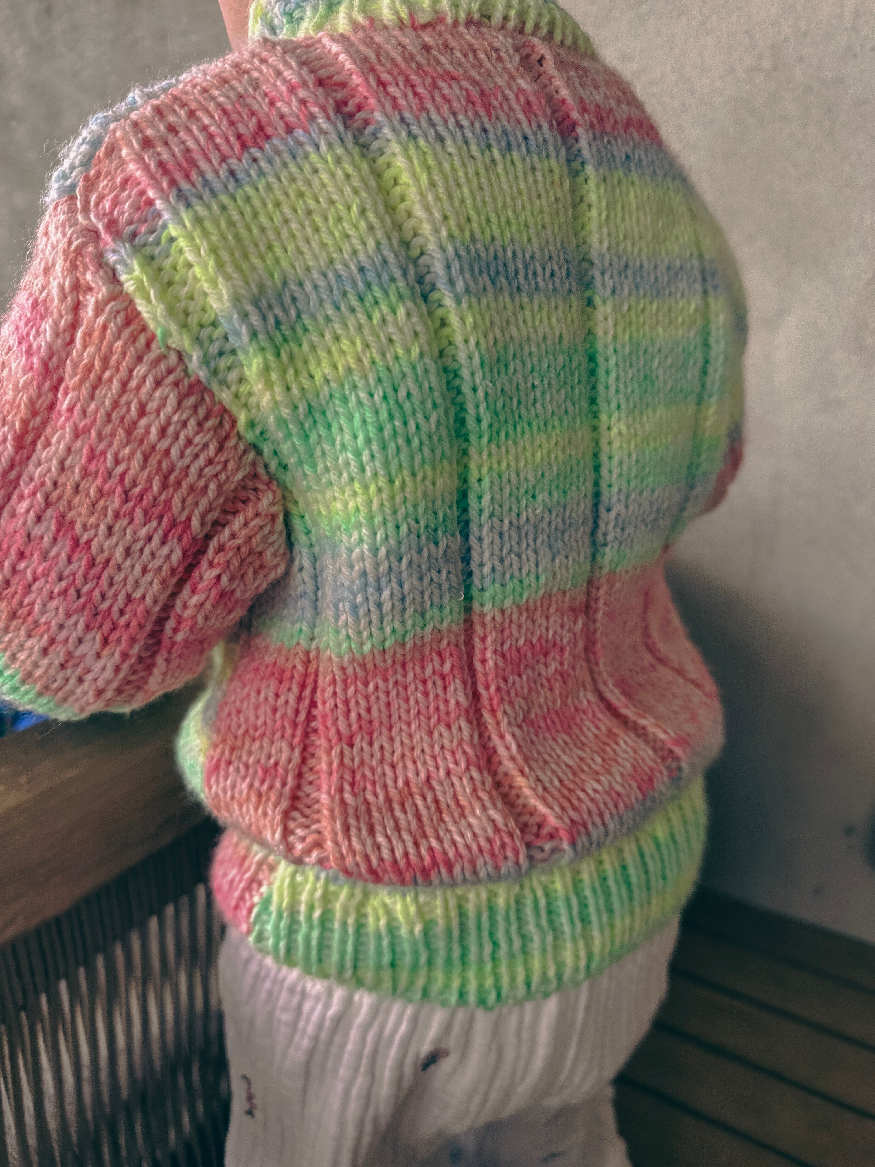 A close-up back view of a child wearing a hand-knit sweater with horizontal stripes in pastel pink, green, yellow, and light blue, paired with white knit pants.