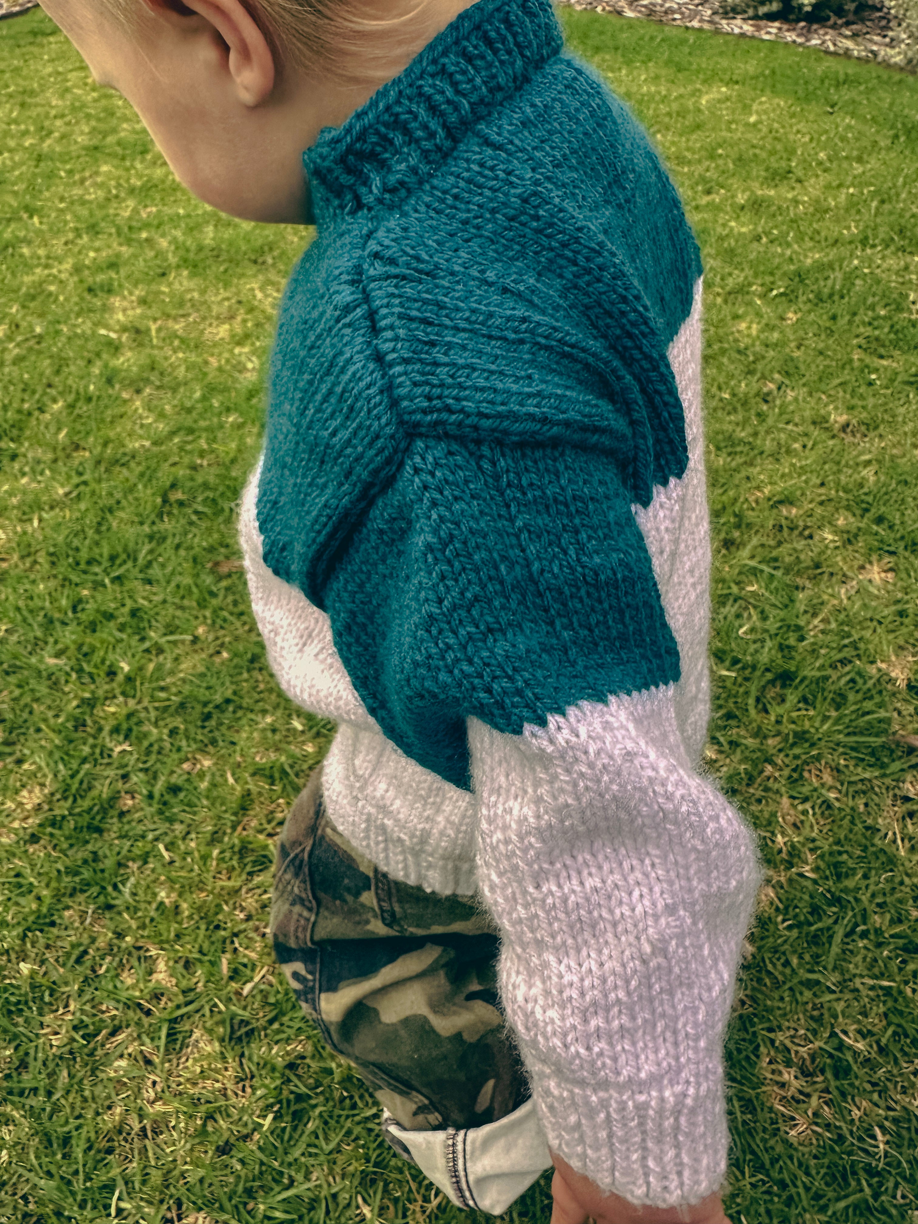 A close-up side view of a child outdoors on grass, wearing a hand-knit sweater with a dark teal mock neck and upper body, and white sleeves, paired with camouflage pants.