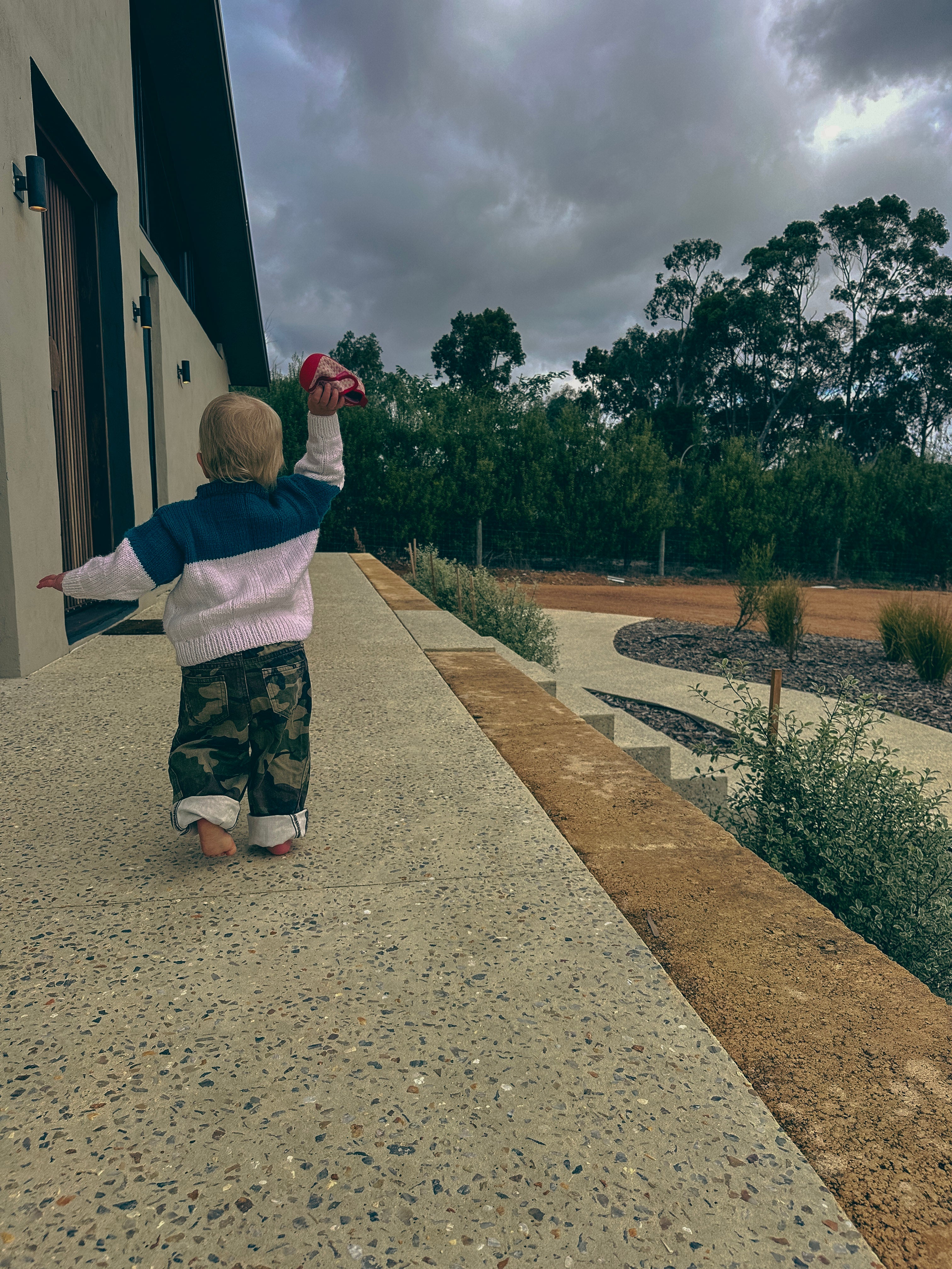 A toddler stands on a concrete walkway outdoors, facing away from the camera and holding a red cup high in the air. The child is wearing a navy and white handknitted sweater with camouflage pants.