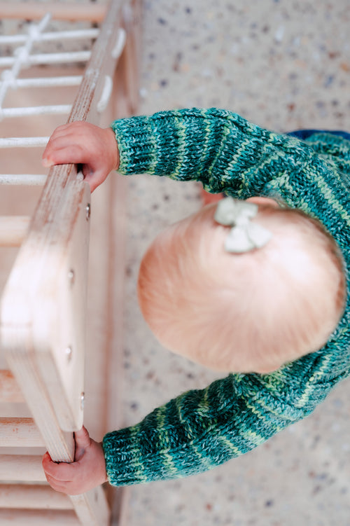 Child wearing a green and blue striped sweater holding the edge of a wood cot