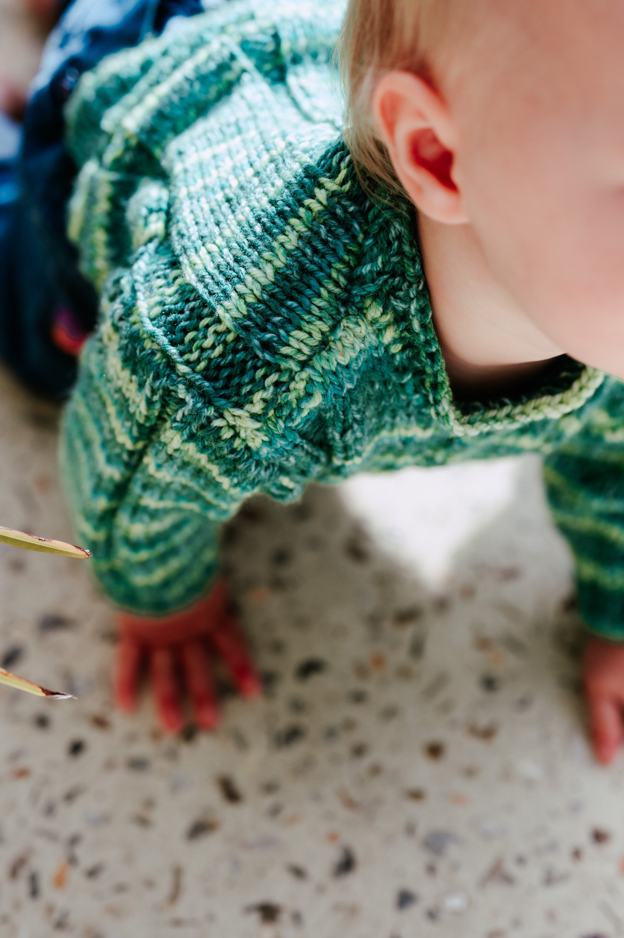 Baby wearing a green knitted sweater crawling on a speckled floor.