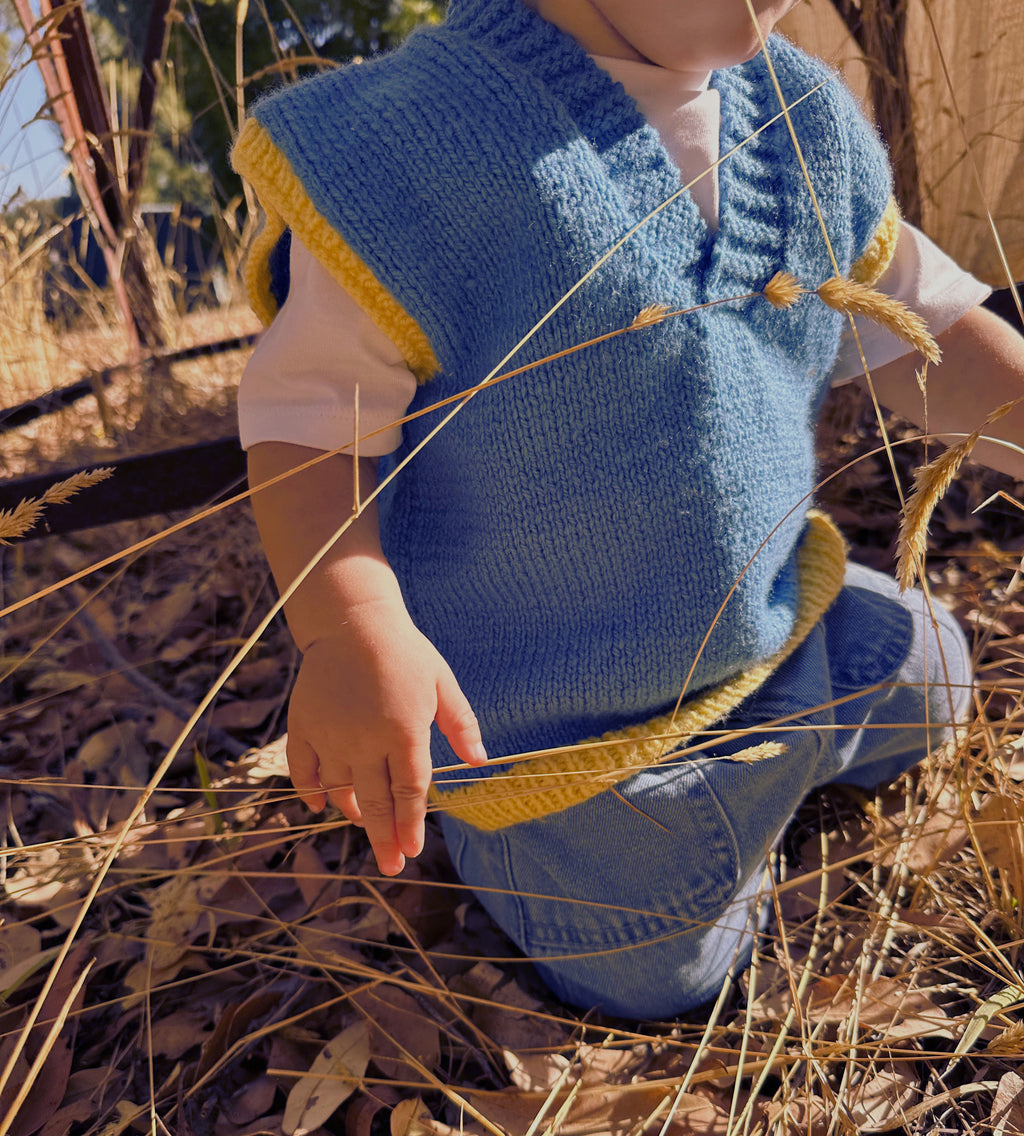 Child wearing a blue knitted vest with yellow trim kneeling in tall grass.