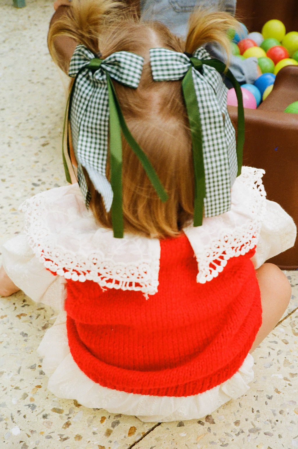 Child wearing a red sweater vest with white lace trim and green bows in hair, sitting on a concrete textured surface next to a ball pit.