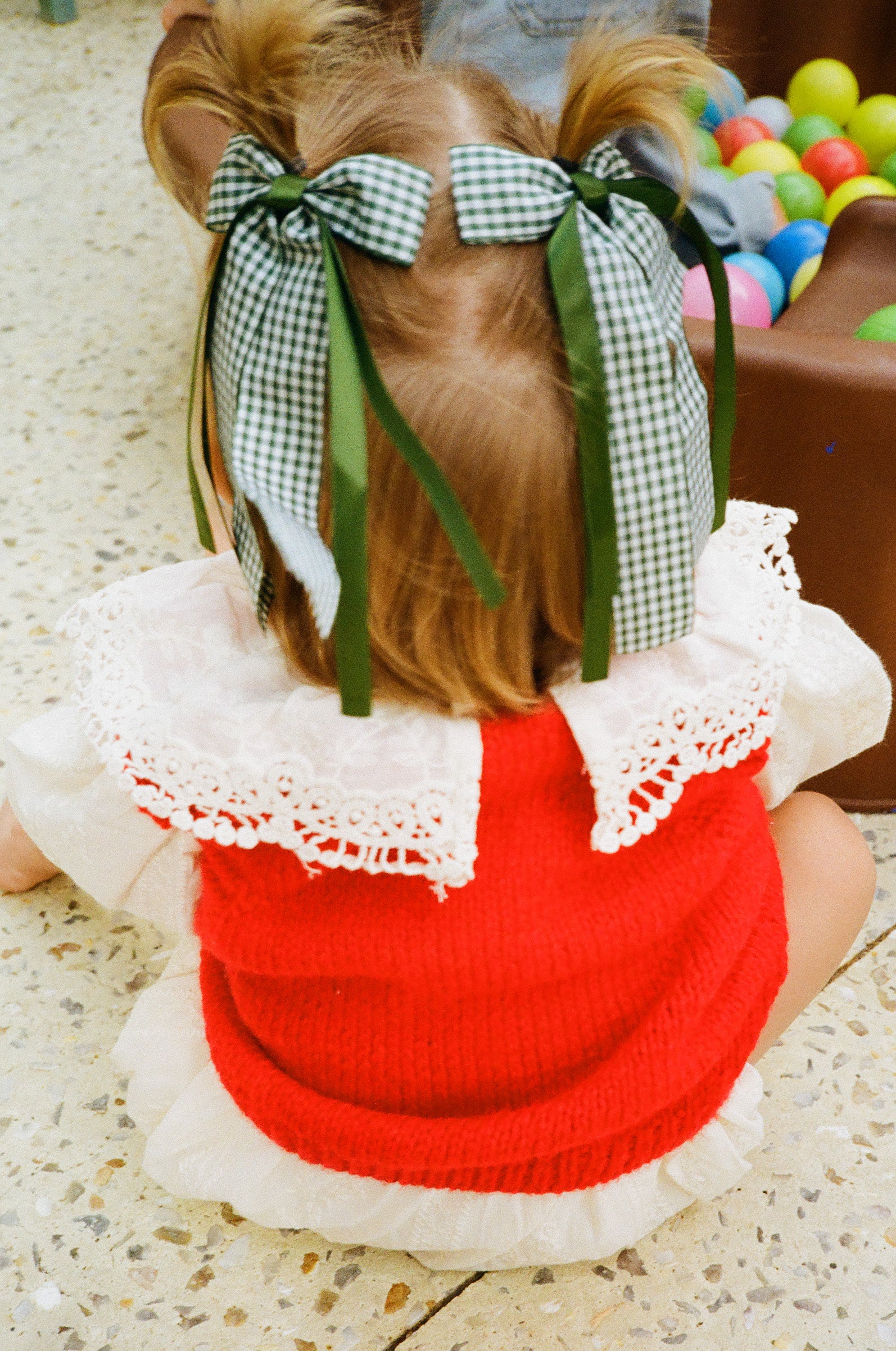 Child wearing a red sweater vest with white lace trim and green bows in hair, sitting on a concrete textured surface next to a ball pit.