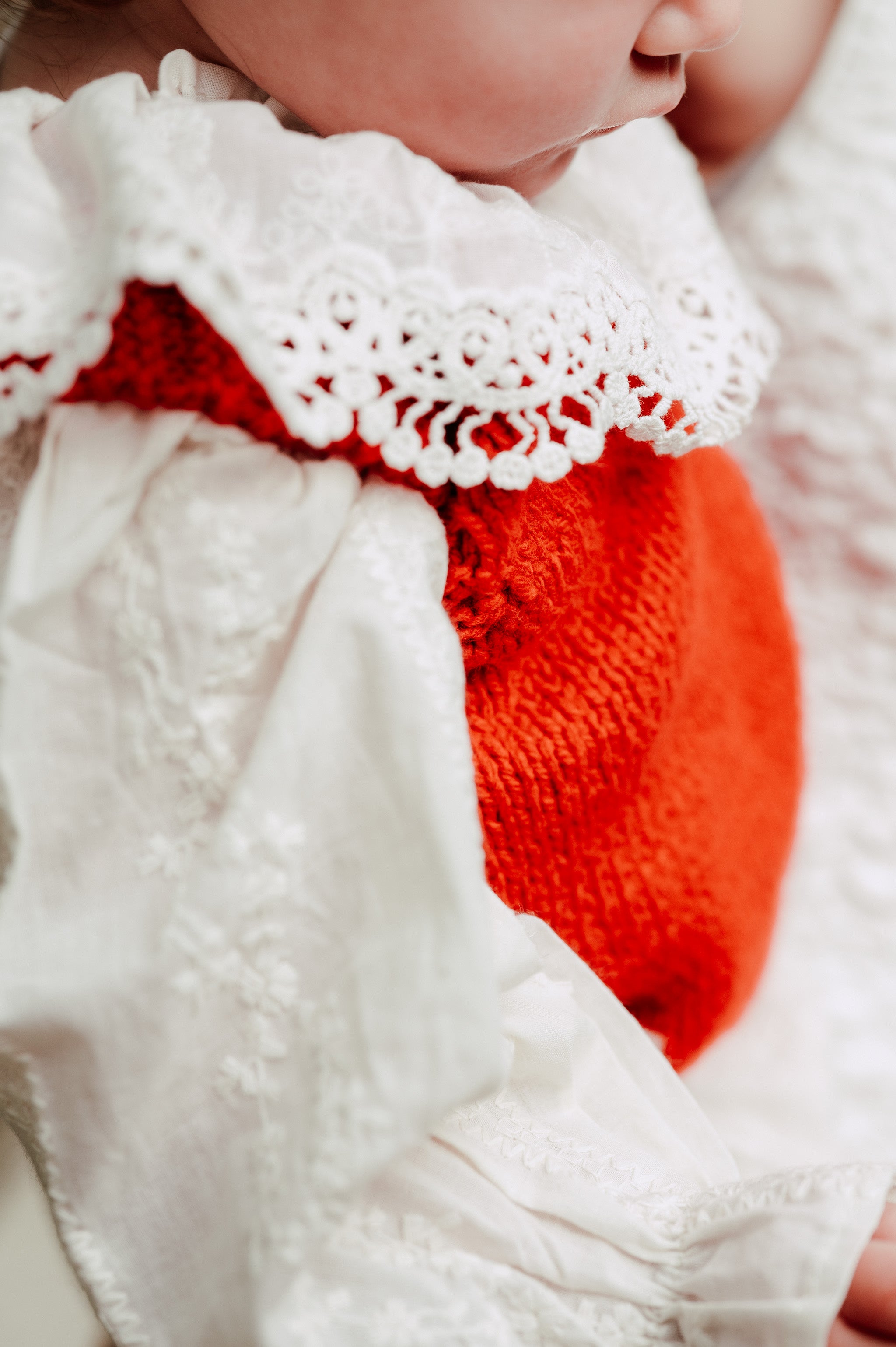 Close-up of a baby wearing a red and white knitted outfit with lace trim.