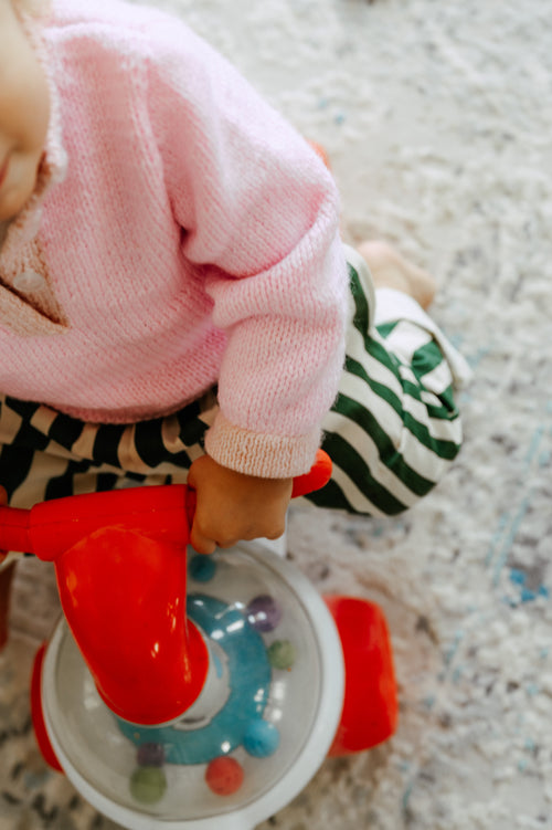Child in a pink sweater and green striped pants, sitting on a red toy scooter