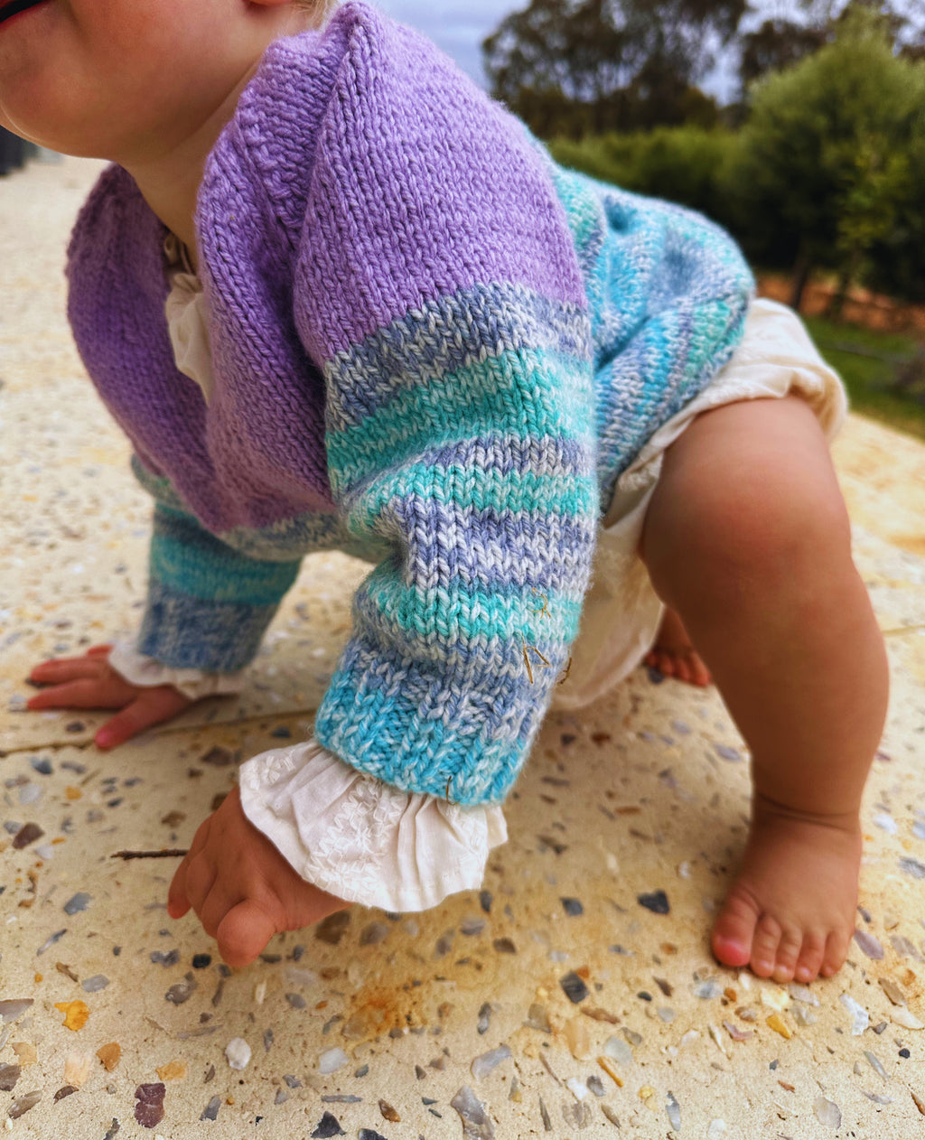 Baby wearing a colorful knitted outfit on a textured surface with greenery in the background