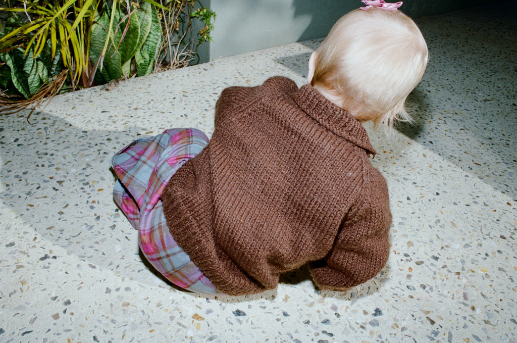Child in a brown sweater and plaid pants crawling on a concrete surface.
