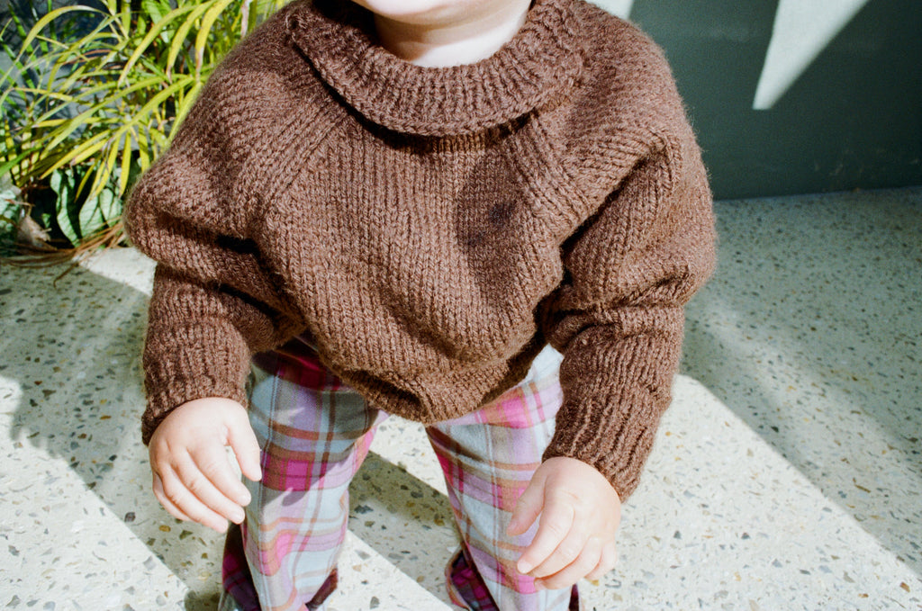 Child wearing a brown knitted sweater and plaid pants outdoors.
