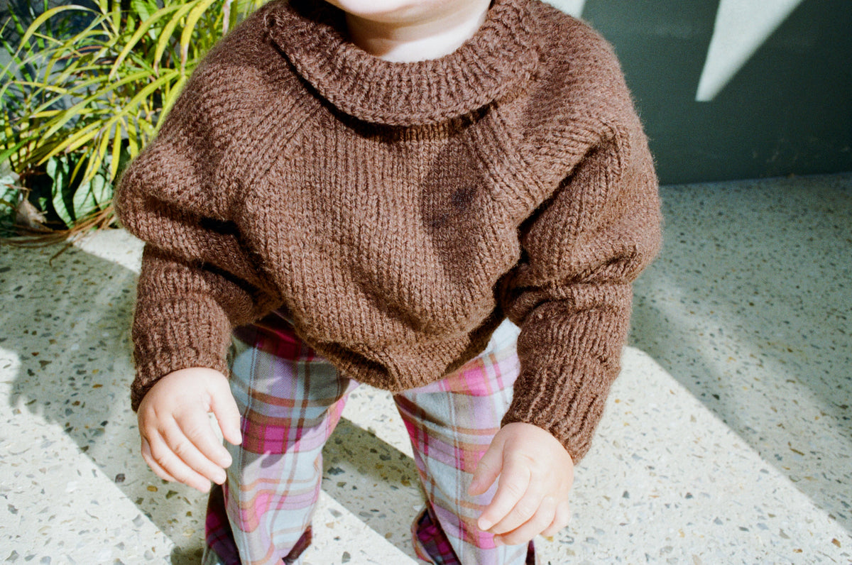 Child wearing a brown knitted sweater and plaid pants outdoors.