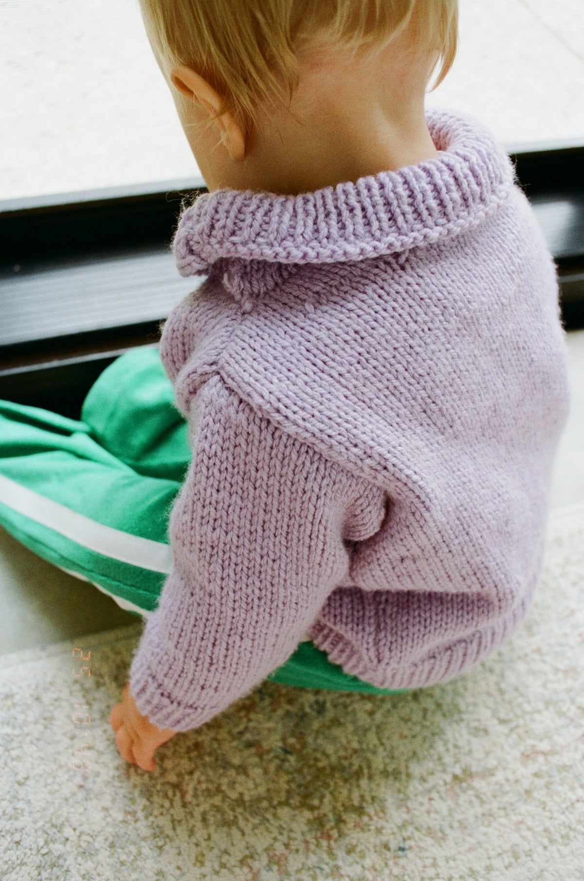 Child wearing a purple knitted sweater and green pants on a carpeted floor.