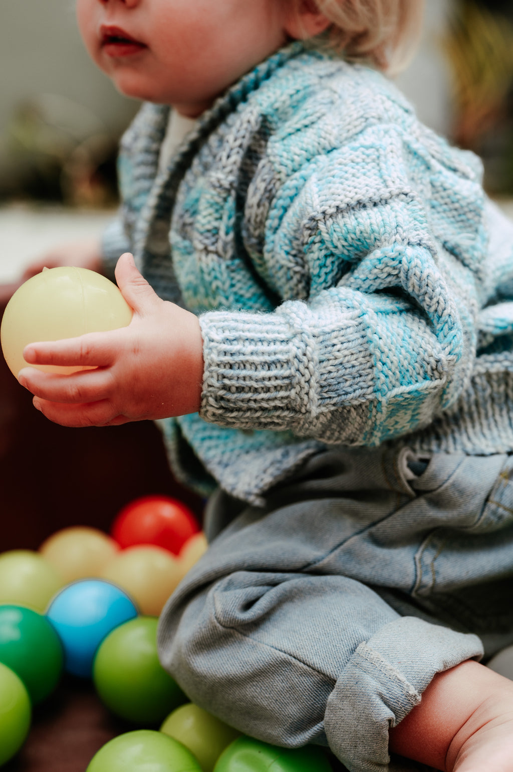 Child in a light blue and grey knitted striped cardigan holding a ball in a colourful ball pit
